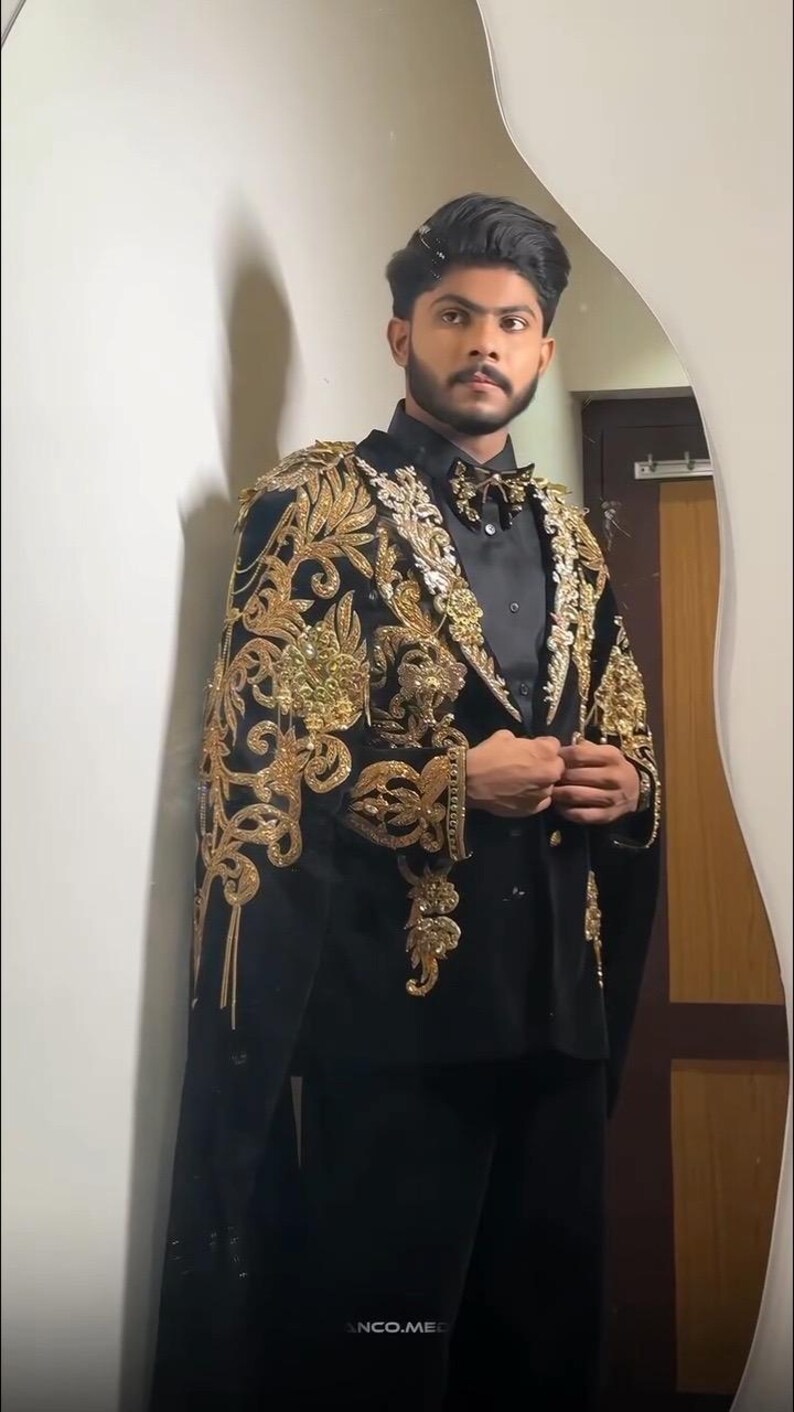 May include: Black velvet suit with gold embroidered detailing. The suit features a cape-like design with intricate floral and scrollwork patterns. The ensemble includes a black shirt and bow tie, creating a formal and elegant look.