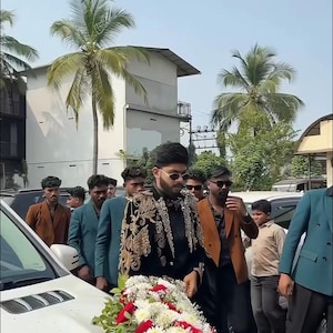 May include: A group of people walking near a white car decorated with a floral arrangement of red and white flowers. The person in the front is wearing a black jacket with gold embroidery. The text "MANCO.MEDIA" is visible.