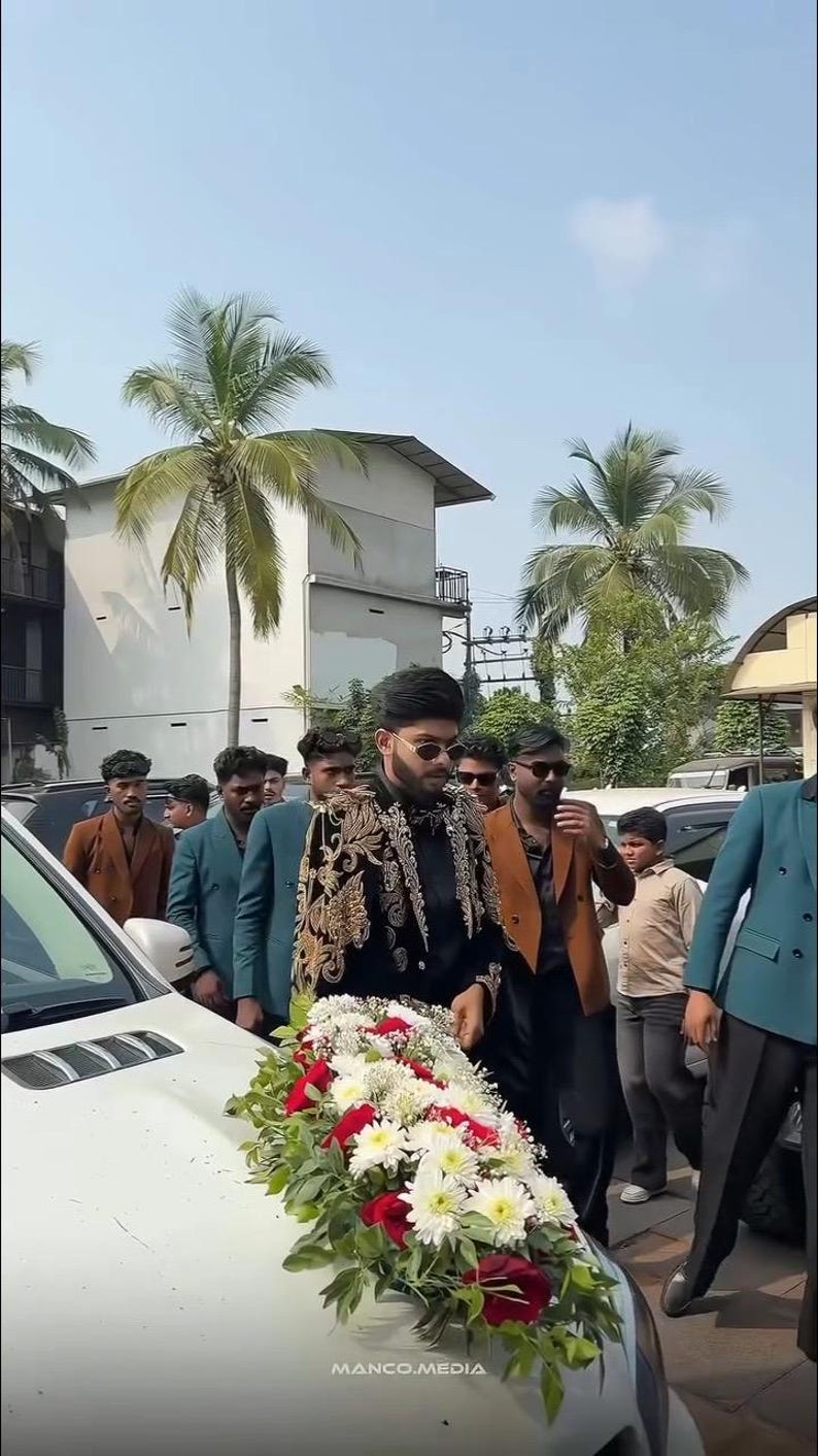 May include: A group of people walking near a white car decorated with a floral arrangement of red and white flowers. The person in the front is wearing a black jacket with gold embroidery. The text "MANCO.MEDIA" is visible.
