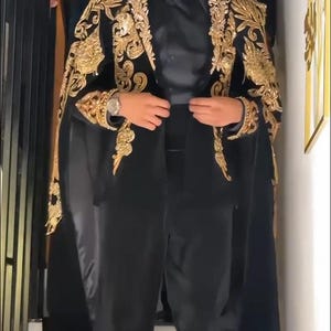 May include: A black suit with gold embroidered detailing on the shoulders and sleeves. The suit includes a cape-like overlay and is paired with a black shirt and pants. The image features a person wearing sunglasses and a watch.