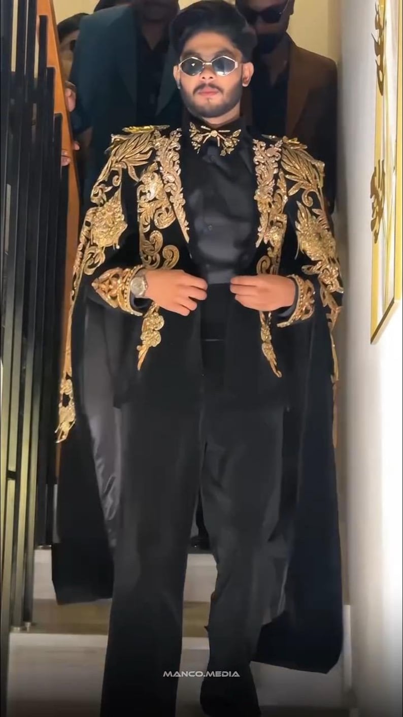 May include: A black suit with gold embroidered detailing on the shoulders and sleeves. The suit includes a cape-like overlay and is paired with a black shirt and pants. The image features a person wearing sunglasses and a watch.