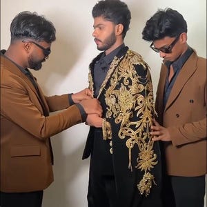 May include: Three people are shown, two on either side of a person wearing a black outfit with gold embroidery. The person on the left is wearing a brown blazer, and the person on the right is wearing a similar brown blazer. The text "MANCO.MEDIA" is at the bottom.