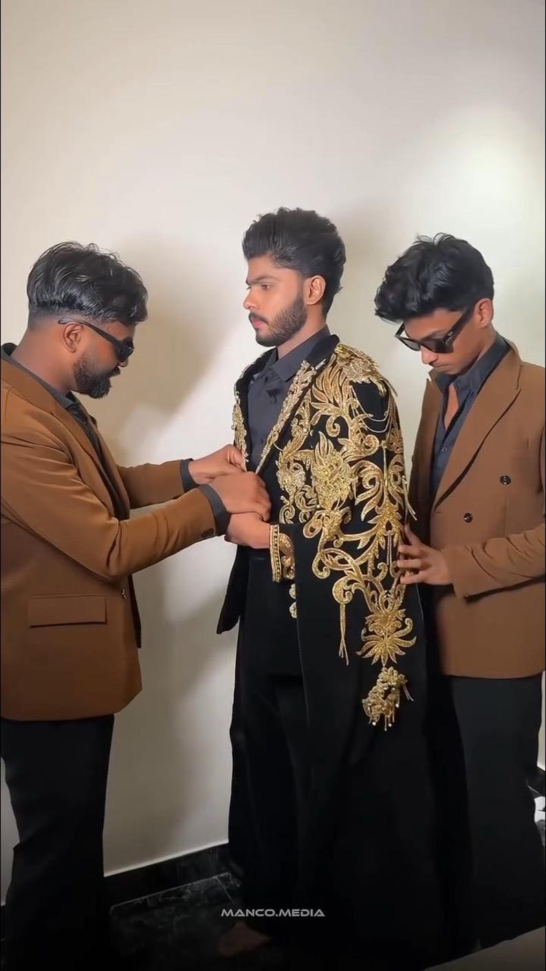 May include: Three people are shown, two on either side of a person wearing a black outfit with gold embroidery. The person on the left is wearing a brown blazer, and the person on the right is wearing a similar brown blazer. The text "MANCO.MEDIA" is at the bottom.