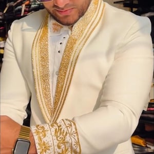 May include: Ivory suit with gold embroidered detailing. The suit features gold accents on the lapels, cuffs, and collar. The person is wearing a gold watch and sunglasses. The suit is a formal style, suitable for special occasions.