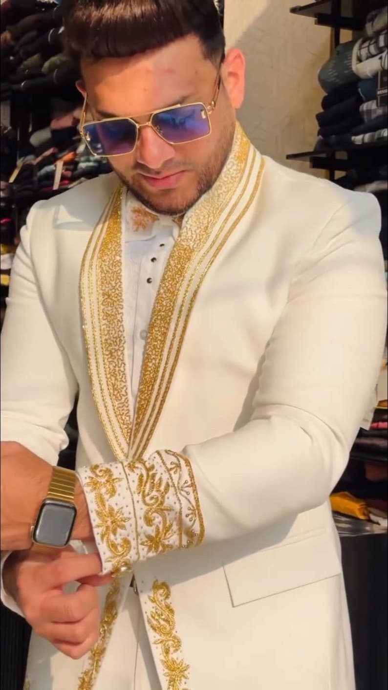 May include: Ivory suit with gold embroidered detailing. The suit features gold accents on the lapels, cuffs, and collar. The person is wearing a gold watch and sunglasses. The suit is a formal style, suitable for special occasions.