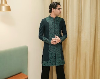 Peut inclure: Longue veste vert foncé avec broderies vertes complexes, portée avec un pantalon noir. La veste a un col mandarin et une coupe ajustée. La tenue est complétée par des chaussures noires. La veste mesure environ 100 cm de long.
