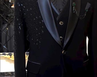 May include: A formal black tuxedo with a satin lapel, bow tie, and vest. The jacket features intricate beaded embellishments on the shoulder. The ensemble includes a black dress shirt and a decorative chain with a green gem.
