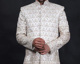 May include: Ivory-colored, long-sleeved jacket with intricate gold embroidery. The jacket features a mandarin collar and ornate patterns across the chest, sleeves, and hem. The garment is worn over white pants, creating a formal ensemble.