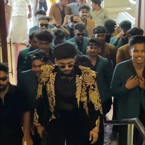May include: A group of people walking down stairs. The person in front is wearing a black outfit with gold embroidery on the jacket. Other people are wearing suits and sunglasses. The text "MANCO.MEDIA" is visible.