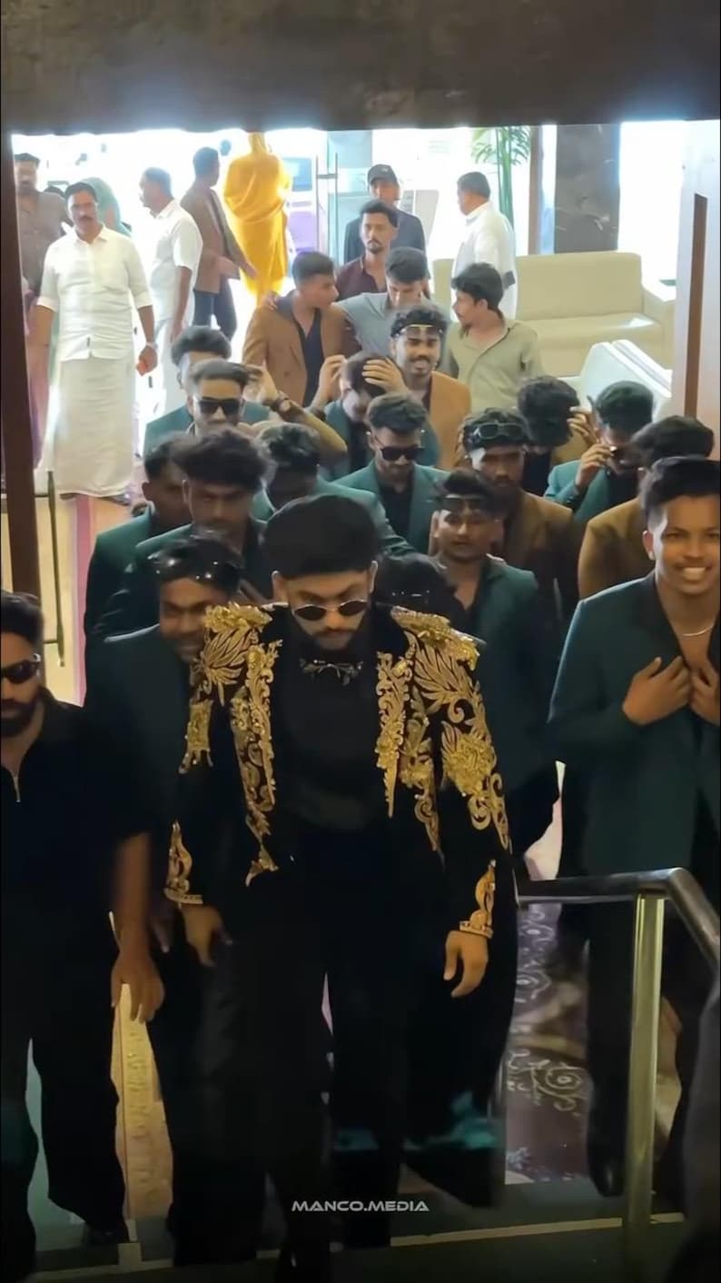 May include: A group of people walking down stairs. The person in front is wearing a black outfit with gold embroidery on the jacket. Other people are wearing suits and sunglasses. The text "MANCO.MEDIA" is visible.