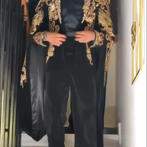May include: A black suit with gold embroidery on the shoulders and sleeves. The suit includes a cape-like design and is paired with black pants. The person is wearing sunglasses and a bow tie. The image is taken indoors.