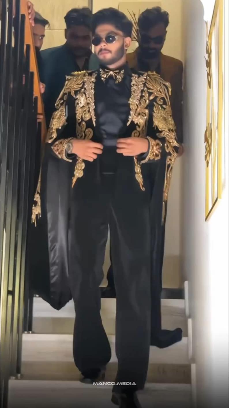 May include: A black suit with gold embroidery on the shoulders and sleeves. The suit includes a cape-like design and is paired with black pants. The person is wearing sunglasses and a bow tie. The image is taken indoors.