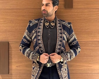 Puede incluir: Un blazer azul marino con intrincados bordados dorados, combinado con pantalones a juego y una camisa negra. El blazer presenta patrones detallados en los bordes y las mangas. Un reloj dorado y un cinturón con hebilla dorada completan el conjunto.