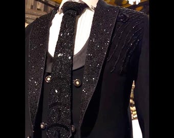 May include: A formal black suit with a white dress shirt and black tie. The suit jacket and tie are embellished with sparkling black beads. The jacket has a notched lapel and gold buttons. The suit is displayed on a mannequin.