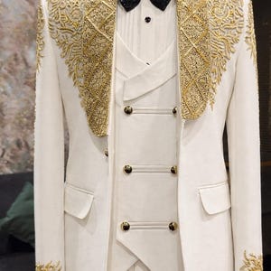 Può includere: Abito formale color avorio con ricami dorati intricati su revers, polsini e spalle. L'abito include un gilet con bottoni dorati e un papillon nero con paillettes. Un ensemble sofisticato per occasioni speciali.