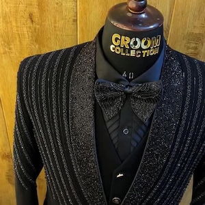 Può includere: Completo da smoking nero con abbellimenti di perline, esposto su un manichino. Il completo include camicia nera, gilet e papillon. Le parole "GROOM COLLECTION" sono visibili sul collo del manichino.