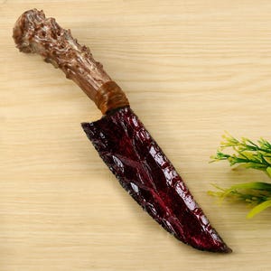Puede incluir: Un cuchillo decorativo con una hoja dentada de color rojo oscuro, translúcida y con textura. El mango está hecho de un material marrón nudoso que recuerda a una cornamenta, sujeto a la hoja con un cordón marrón enrollado en la guarda.