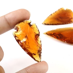 May include: Close-up of several amber-colored, translucent arrowhead-shaped pieces. One is held between fingers, while two others are in the background. The pieces have a rough, textured appearance and are set against a white background.