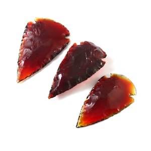 May include: Three translucent, reddish-brown arrowhead-shaped objects are arranged on a white background. The edges of the objects have a slightly darker hue, and the overall appearance suggests a glassy or polished texture.