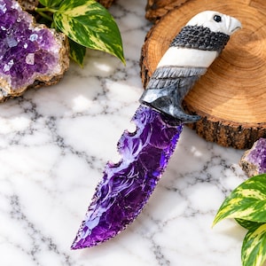May include: A decorative knife featuring a translucent, deep purple, jagged-edged blade made of stone. The handle is intricately carved in the shape of a white and gray eagle head with detailed feather textures and a dark, sharp beak.