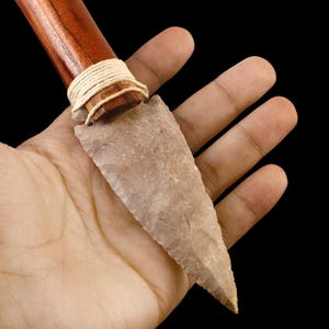 Puede incluir: Una punta de lanza primitiva, de mano, con una hoja de piedra y un mango de madera. La hoja es de color marrón claro y el mango está envuelto con un cordón de color natural.