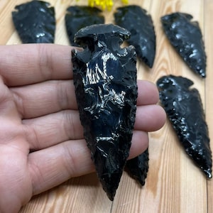 May include: A collection of obsidian arrowheads, each with a dark, glossy finish. The arrowheads are displayed on a wooden surface, with a few in the foreground and others in the background. The edges of the arrowheads are sharp and well-defined.