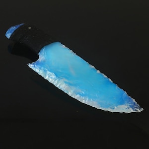 May include: A hand-crafted knife with a black handle and a translucent, light blue blade. The blade has a rough, chipped edge and a polished surface. The knife is set against a black background.