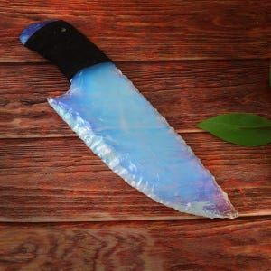 May include: A handcrafted knife with a blade made of opalescent material, displaying a range of blues and whites. The handle is wrapped in black material. The knife is displayed on a wooden surface.