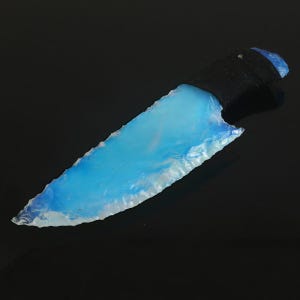 May include: A handcrafted knife with a light blue, translucent blade and a black handle. The blade has a rough, chipped edge, and the handle appears to be wrapped in a textured material. The knife is set against a dark background.
