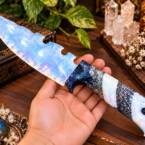 May include: A decorative knife featuring a serrated, iridescent blue blade that mimics the appearance of opal. The handle is crafted with a textured dark blue finish, a diagonal white inlay, and a pommel shaped like an eagle head.