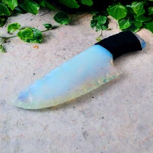 May include: A handcrafted, opalescent knife with a black wrapped handle. The blade has a light blue and white color with a textured edge. The knife is displayed on a textured, light gray surface with green foliage in the background.