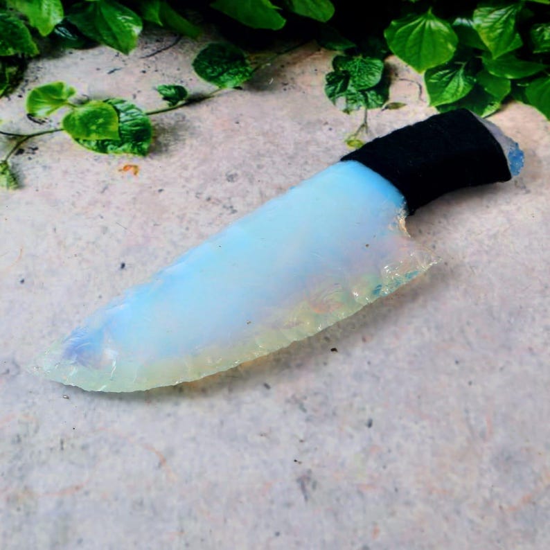 May include: A handcrafted, opalescent knife with a black wrapped handle. The blade has a light blue and white color with a textured edge. The knife is displayed on a textured, light gray surface with green foliage in the background.