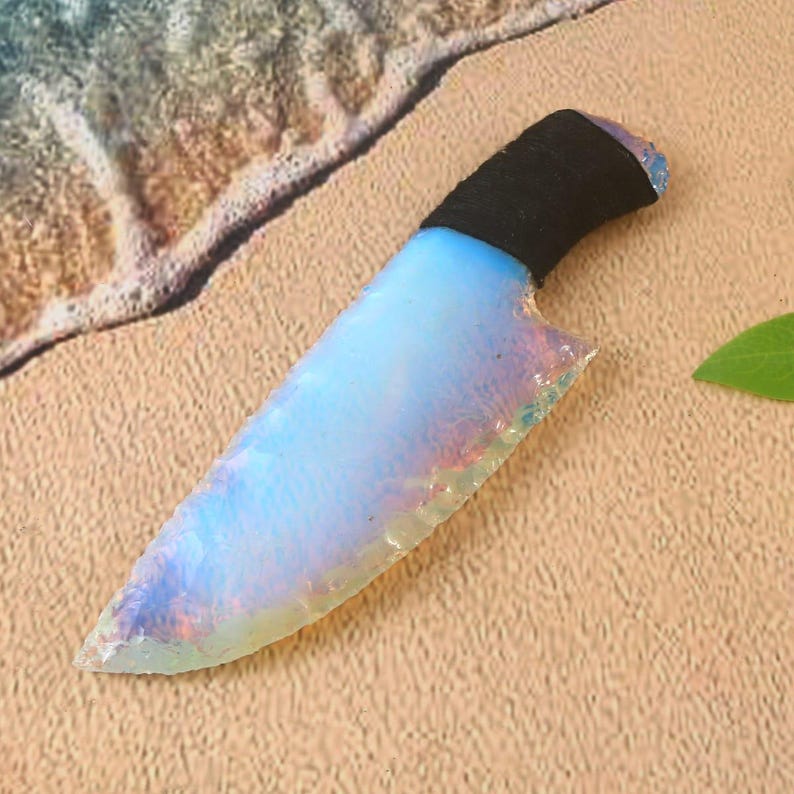May include: A decorative knife with a blade that has a pearlescent, opalescent appearance. The handle is wrapped in black material. The knife is resting on a sandy surface.