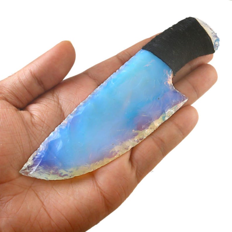 May include: A handcrafted knife with a blade of opalescent blue and white hues. The handle is wrapped in black material. The knife is held in a hand against a white background.