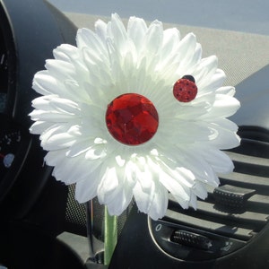 May include: A white daisy car vent clip with a large red gem center and a small ladybug accent. The flower is made of fabric and is attached to a silver clip. The car vent clip is designed to add a decorative touch to a vehicle's interior.
