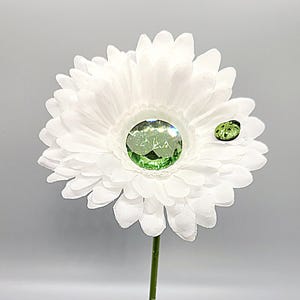 Puede incluir: Una margarita blanca con un strass verde en el centro y una pequeña mariquita verde en un pétalo.
