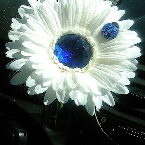 Puede incluir: Una margarita blanca con una gema azul en el centro y una gema azul más pequeña en el lateral. La flor está en un coche.