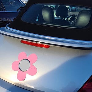 Op de afbeelding: Een zilveren cabriolet met een zwart dak en een roze bloemvormige magneet. De magneet heeft een zilveren midden en is bevestigd aan de kofferbak van de auto. Een rood remlicht is zichtbaar boven de magneet.