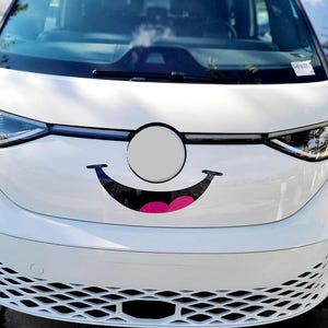 May include: A white van with a large smiling face decal on the front. The smiling face has a black curved mouth and pink lips. The van has a black grill and headlights.