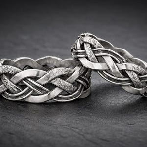 Può includere: Due anelli color argento con un design a nodo celtico. Gli anelli presentano un motivo intrecciato con una finitura strutturata. Sono presentati su una superficie scura e strutturata, che accentua gli intricati dettagli degli anelli.