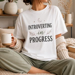 May include: A white short-sleeved t-shirt featuring the black text "INTROVERTING in PROGRESS" centered on the chest. The design includes a small crescent moon, stars, and two leafy branch illustrations flanking the word "in" in green.