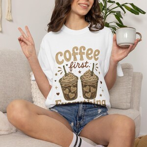 May include: A white t-shirt featuring a graphic print of two iced coffee drinks with whipped cream and heart shapes. The text reads "coffee first" above the drinks and "Everything" below them, accented with small stars and hearts in brown.