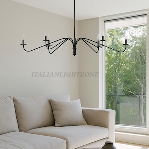 Op de afbeelding: Een zwarte, moderne kroonluchter met meerdere gebogen armen, elk met een gloeilamp. De kroonluchter hangt in een woonkamer met een beige bank en een houten salontafel. De tekst "ITALIANLIGHTZONE" is zichtbaar.