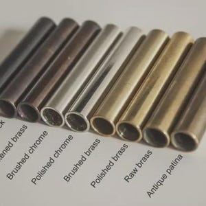 Pode incluir: Uma cole&ccedil;&atilde;o de tubos de metal em v&aacute;rios acabamentos, incluindo preto fosco, preto antigo e cromo polido. A imagem exibe uma gama de cores do escuro ao claro, com etiquetas que identificam cada acabamento. Os tubos est&atilde;o dispostos diagonalmente.