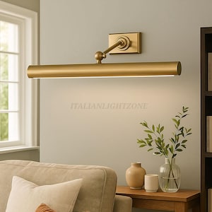 May include: A gold-toned picture light fixture mounted on a light gray wall. The fixture has a rectangular base, an adjustable arm, and a long, cylindrical light source. The text "ITALIANLIGHTZONE" is visible. The scene includes a sofa, side table, and decorative items.