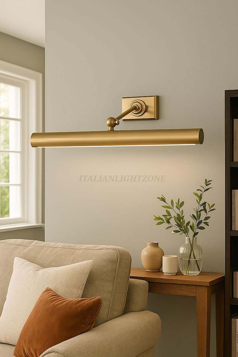 May include: A gold-toned picture light fixture mounted on a light gray wall. The fixture has a rectangular base, an adjustable arm, and a long, cylindrical light source. The text "ITALIANLIGHTZONE" is visible. The scene includes a sofa, side table, and decorative items.
