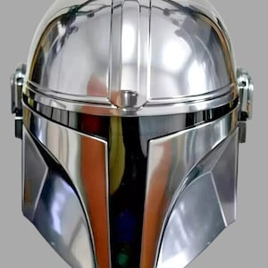 Peut inclure: Casque argenté brillant de style Mandalorien, avec une visière en forme de T. Le casque présente un sommet arrondi avec des arêtes verticales et une finition métallique. La visière est noire et réfléchissante.