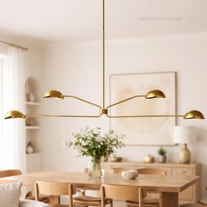 Pode incluir: Um candelabro moderno, dourado, com cinco luzes em forma de c&uacute;pula. A lumin&aacute;ria est&aacute; suspensa do teto e tem um design minimalista. A imagem tamb&eacute;m mostra uma mesa de jantar de madeira, cadeiras e um vaso de flores, sugerindo uma sala de jantar.