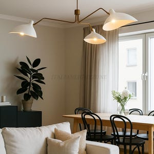 May include: A contemporary chandelier with a brass frame and white lampshades. The light fitting hangs above a dining table and chairs. The text "ITALIANLIGHTZONE" is visible. The room is decorated in neutral tones.