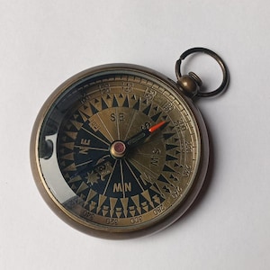 May include: A vintage brass compass with a clear glass cover. The compass face features directional markings and degree indicators. A red needle points north. The compass has a small ring for attachment.